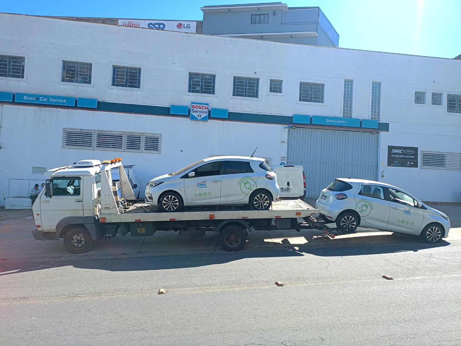 Veículo branco sobre a plataforma de um caminhão de guincho em frente a um prédio.
