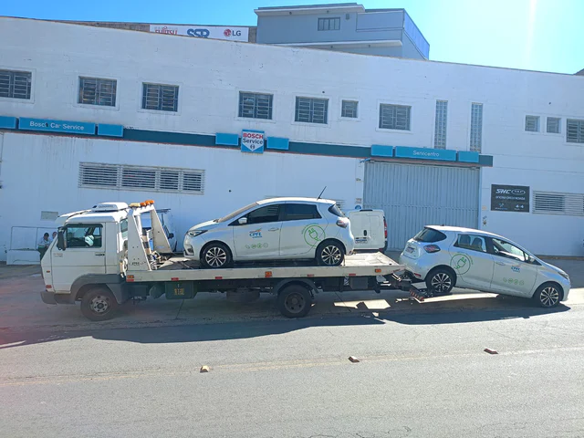 Veículo branco sobre a plataforma de um caminhão de guincho em frente a um prédio.