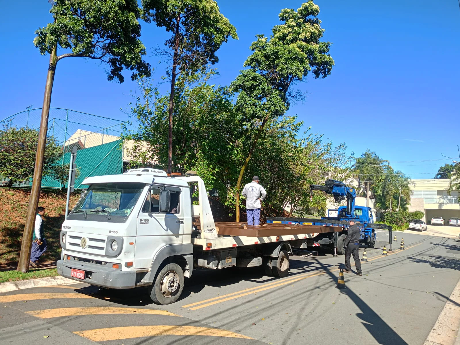 Caminhão-plataforma branco em operação ao lado de um caminhão com guindaste.